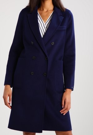 Kobieta w podwójnym granatowym płaszczu na białej sukience z czarnymi pionowymi paskami, stojąca na jednolitym tle.