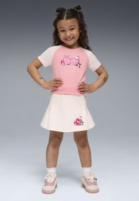 Uśmiechnięta dziewczynka z ciemnymi włosami stoi z rękami na biodrach, ubrana w różowo-białe ubranie z Hello Kitty, do którego pasują różowe trampki i tymczasowe tatuaże.