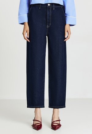 Donna che indossa jeans a gamba larga blu scuro, camicia a righe blu e tacchi a punta rossi con cinturini alla caviglia, in piedi contro uno sfondo neutro.
