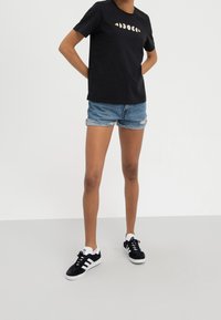 Zwarte katoenen t-shirt met korte mouwen, versierd met meerkleurige madeliefjes borduurwerk over de borst; gedragen met versleten denim shorts en zwarte sneakers.