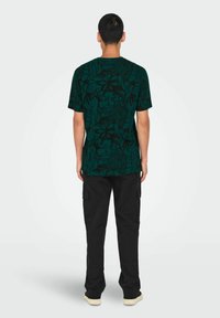Camiseta verde de manga corta con un patrón floral negro, combinada con pantalones cargo negros. La camiseta tiene un cuello redondo y un corte holgado.