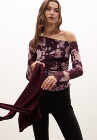 Bordový kvetinový top s dlhým rukávom a off-shoulder dizajnom; priehľadné rukávy, priliehavé telo a štruktúrovaná látka v kombinácii s čiernymi priliehavými nohavicami.