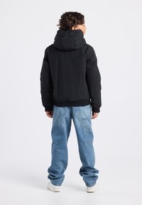 Black padded jacket with a large hood, ribbed hem, and cuffs, paired with loose-fitting light blue jeans and white sneakers.