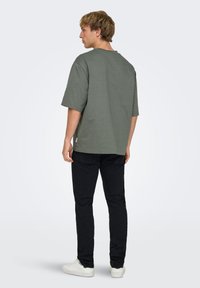 T-shirt gris en coton à manches courtes, coupe décontractée, col rond et étiquette latérale, porté avec un pantalon noir et des baskets blanches.