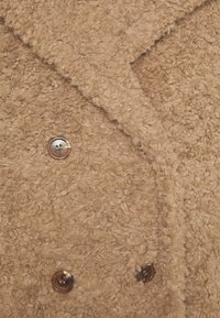 Texture corta e riccia di lana in tessuto color tostato con tre bottoni marrone scuro e un colletto piegato su un cappotto o giacca.