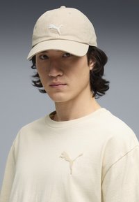 Casquette de baseball beige avec un logo brodé blanc, portée avec un t-shirt beige clair assorti comportant un logo en relief sur la poitrine.