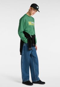 Grünes Langarmshirt mit gelbem Grafikdesign, schwarzer Pullover um die Taille gebunden, weite blaue Jeans und schwarze Sneakers, ergänzt durch eine schwarze Mütze.