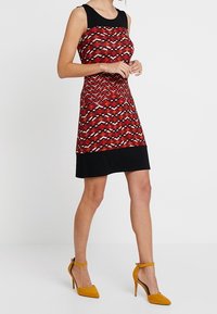 Robe rouge et noire avec un motif géométrique, sans manches, col rond, et panneau inférieur noir. Assortie à des chaussures à talons jaunes.