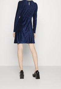 Abito in velluto blu navy con pieghe verticali, maniche lunghe e dettaglio con apertura a chiave sulla parte posteriore. Abbinato a scarpe con tacco a blocco nere.