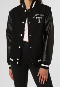 Černá univerzitní bunda s koženými rukávy, bílými knoflíky a vyšívaným logem "T" a "TOMMY JEANS". Hladké manžety a límec s bílými akcenty.