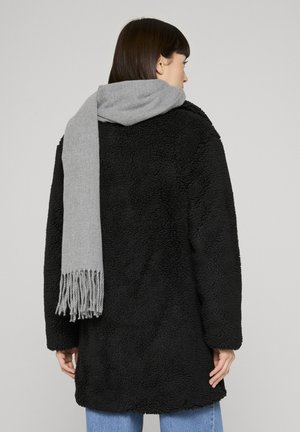 Femme portant un manteau noir à texture et une longue écharpe grise drapée sur son épaule, debout devant un fond clair uni.