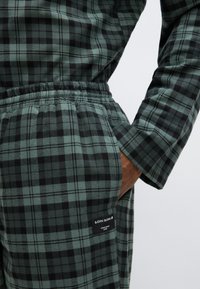 Completo da soggiorno a quadri verdi e neri con vestibilità rilassata, vita elastica e tasche laterali. Il materiale appare morbido con una finitura testurizzata.