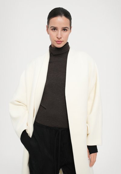 Ivory oversized coat made of soft material, paired with a dark brown turtleneck. Features wide sleeves and a relaxed fit.