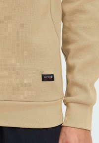 Felpa beige con texture, caratterizzata da un design a coste, patch logo ricamato sul lato sinistro in basso e polsini e orlo a costine.