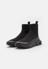 Bottines noires avec un haut extensible et un col texturé semblable à une chaussette. Présente une semelle noire épaisse avec des découpes géométriques et un marquage.