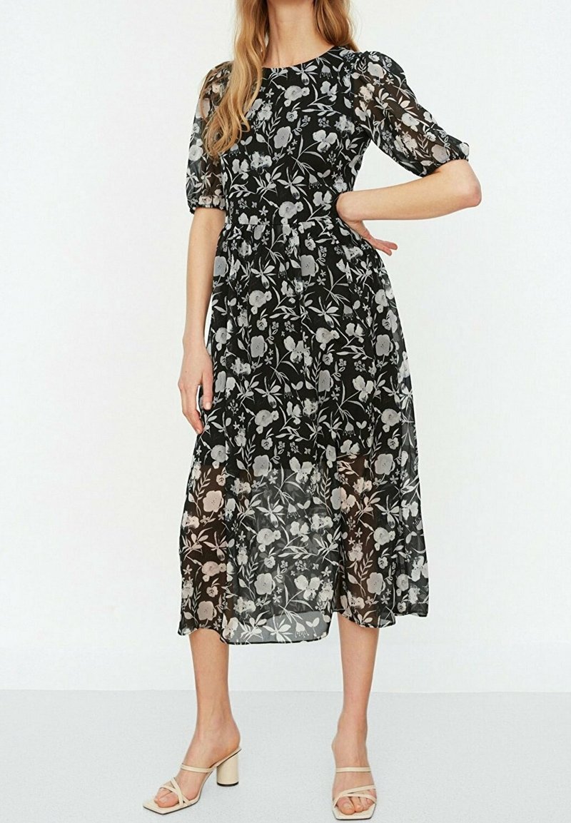 Femme portant une robe midi noire à imprimé floral blanc et manches bouffantes transparentes, associée à des sandales beiges à talons blocs et orteils ouverts.