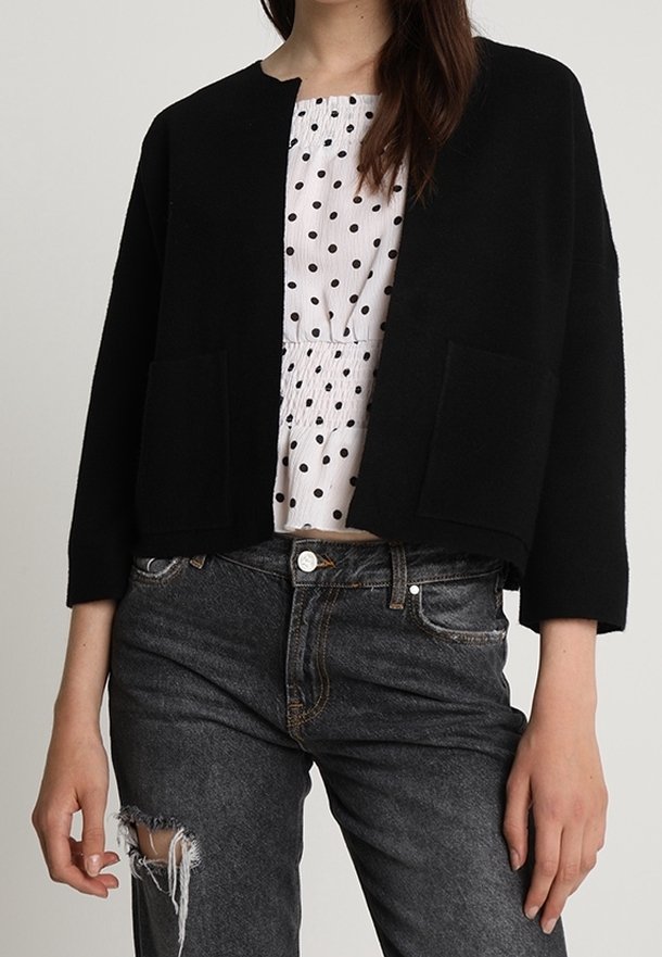 Cardigan noir court à manches longues, avec deux poches avant et une texture douce, associé à une blouse blanche à pois et à un jean gris déchiré.