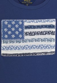 Camiseta azul marino con un diseño de bandera estilo patchwork que presenta patrones florales en azul y blanco, detalles de costura y la etiqueta "Ralph Lauren" en la parte inferior.