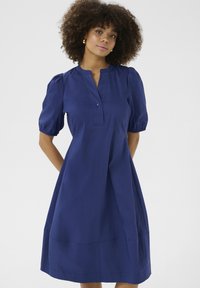 Tamsiai mėlyna suknelė su trumpomis pūkuotomis rankovėmis, mažomis mygtukais suknelėmis ir laisva sijonų dalimi, besitęsiančia iki vidurinių blauzdų. Lygus audinio paviršius.