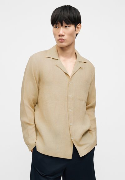 Chemise en lin beige avec col légèrement ouvert, manches longues et poche avant. Présente une coupe décontractée et une fermeture à boutons visible.