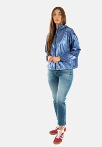 Blauwe glanzende, lichte jas met een capuchon, rits aan de voorkant en elastische manchetten, gecombineerd met strakke jeans en meerkleurige sneakers.