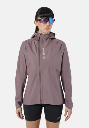 Vrouw draagt een mauve capuchonjack met voorrits en zwarte fietsshorts, met daarnaast gespiegelde sportzonnebril, kijkend naar voren.