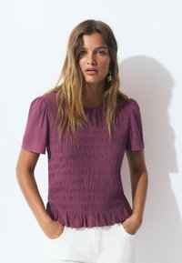Chemisier violet texturé avec des manches courtes bouffantes et un ourlet froncé, porté avec un pantalon blanc. Présente des détails de smocks verticaux.