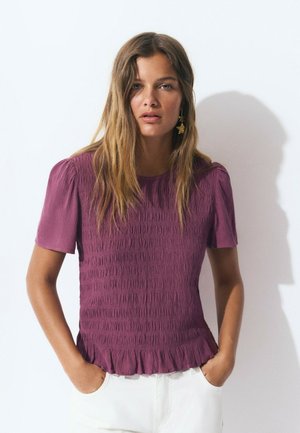 Chemisier violet texturé avec des manches courtes bouffantes et un ourlet froncé, porté avec un pantalon blanc. Présente des détails de smocks verticaux.