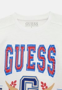 Bílý organický bavlněný tričko s velkým logem "GUESS" v červené a modré barvě, s dalšími dekorativními prvky a akcenty ve zlaté a červené.