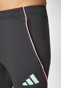 Svart treningbukser med en rosa stripe langs siden, med en lomme og en lyseblå logo på nederste del av benet. Glatt, elastisk stoff.