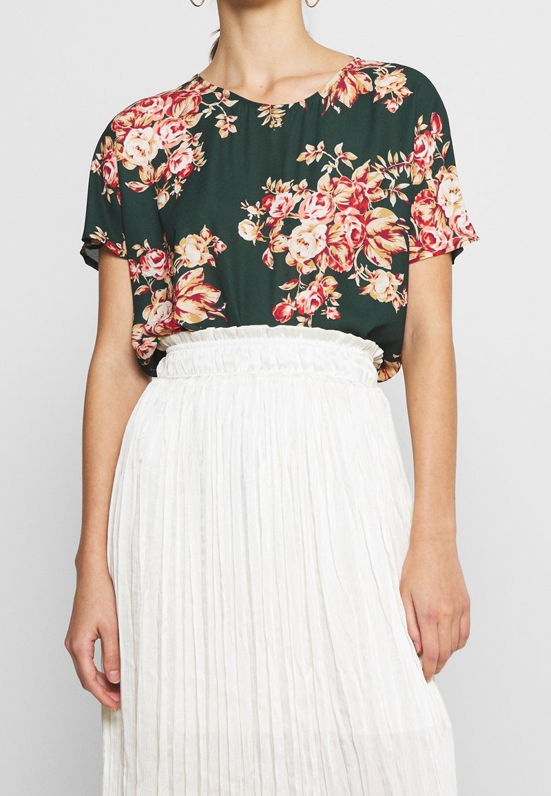 Halenka s krátkým rukávem s tmavě zeleným základem a květinovým vzorem v odstínech růžové a krémové. V kombinaci s bílou skládací sukní.