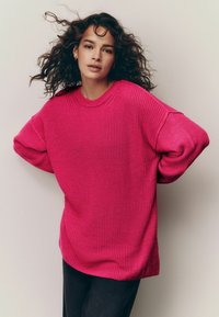 Pulover supradimensionat, de culoare roz intens, tricotat, cu un design texturat, guler rotund cu nervuri și mâneci largi, asortat cu blugi închiși la culoare.