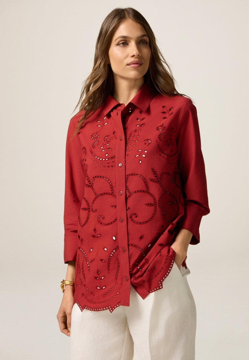 Rotes Hemd mit Knopfleiste, das florale Stickereien und Ausschnittdetails aufweist. Lange Ärmel, abgerundeter Saum und eine glatte Stofftextur.