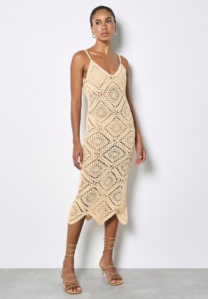 Frau trägt ein ärmelloses beigefarbenes gehäkeltes Midikleid mit Diamantmustern und beigen Riemchenabsätzen, steht vor einer weißen Wand.