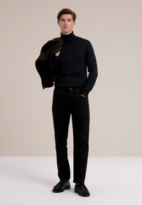 Man in een zwarte coltrui en broek, met een zwart jack over zijn schouder, staande tegen een effen beige achtergrond.