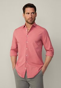 Camisa rosa claro de manga larga con botones, cuello clásico y mangas enrollables. Presenta un pequeño logo bordado en el pecho.