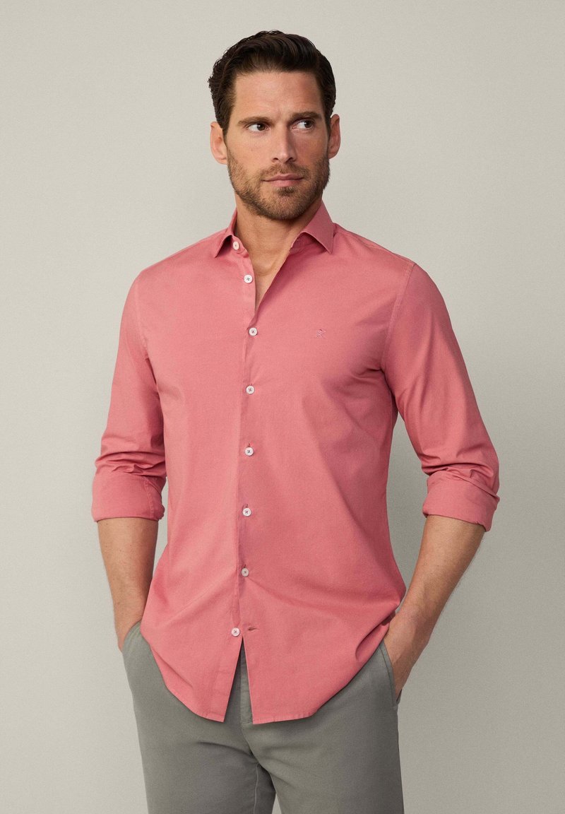 Camisa rosa claro de manga larga con botones, cuello clásico y mangas enrollables. Presenta un pequeño logo bordado en el pecho.