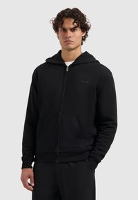 Man in een zwart zip-up hoodie met zakken aan de voorkant en een capuchon, staand met één hand in de zak tegen een effen grijze achtergrond.