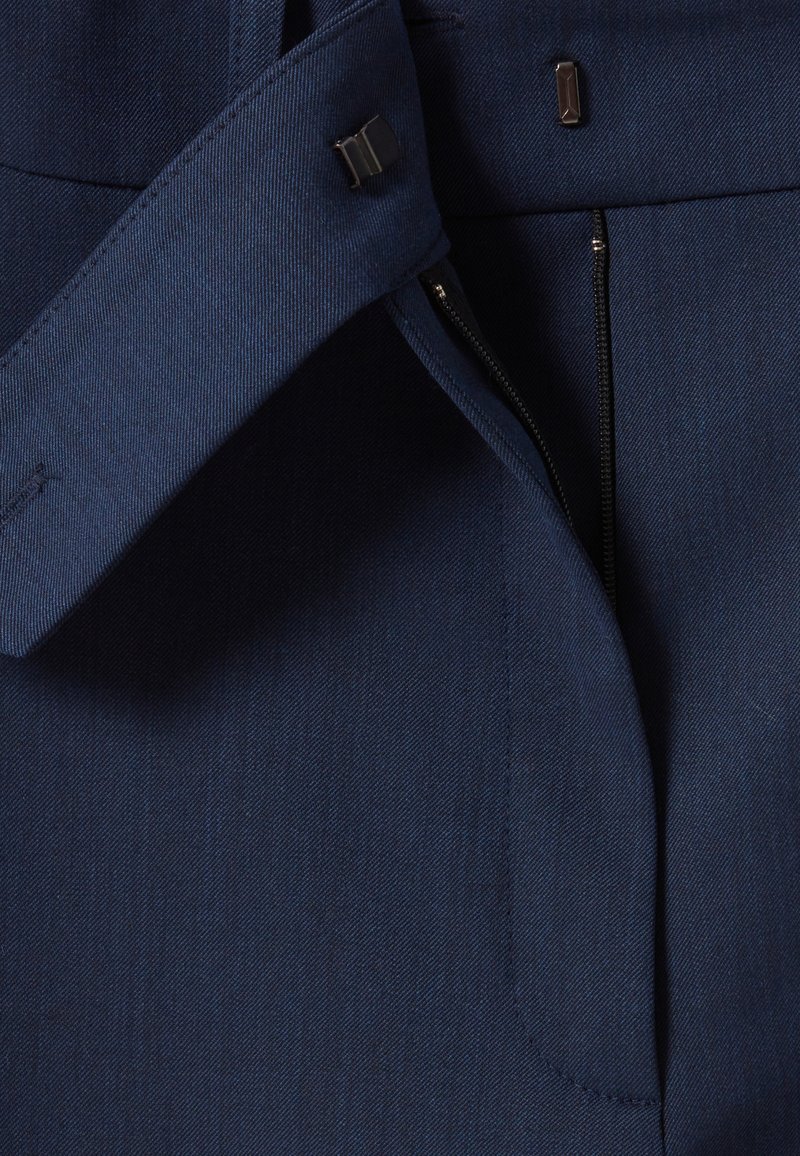 Marineblaue Stoff mit feiner Textur. Verfügt über einen Bund, metallische Akzente und eine mit Reißverschluss verschlossene Öffnung, mit sauberer Naht und tailliertem Design.