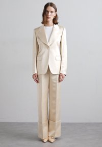 Beige satin kostym med en figursydd blazer med skarpa slag och två framfickor, kombinerad med vida byxor och spetsiga skor.