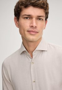 Młody mężczyzna z krótkimi kręconymi brązowymi włosami, ubrany w beżową koszulę z guzikami, patrzący na wprost z neutralnym wyrazem twarzy na jednolitym tle.