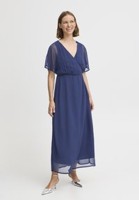Marinblå maxi-klänning i lätt tyg, med korta volangärmar, V-ringning, elastisk midja och ankellång design.