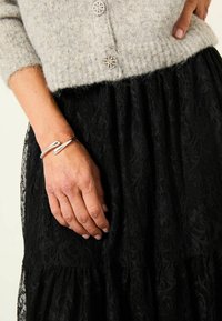 Silbernes Armband an einer Hand mit einem fließenden, wellenartigen Design. Schwarzer Spitzenrock mit floralem Muster und plissierter Textur. Graue Strickjacke darüber.