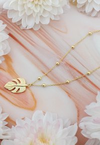 Guld kedja smycke med en monstera blad hänge med utskurna detaljer och runda guld pärlor. Bakgrunden har mjuka rosa och vita blommiga element.