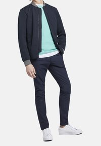 Námořnická bombardovací bunda s šedými akcenty, světlý tyrkysový svetr, texturované tmavé kalhoty a bílé tenisky, vytvářející ležérní outfit.