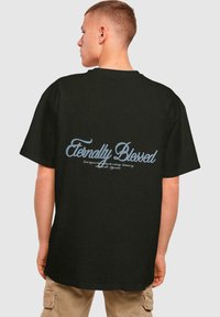 Tricou din bumbac negru cu o croială lejeră, având text albastru cursiv "Eternally Blessed" pe spate. Guler rotund standard și mâneci scurte.