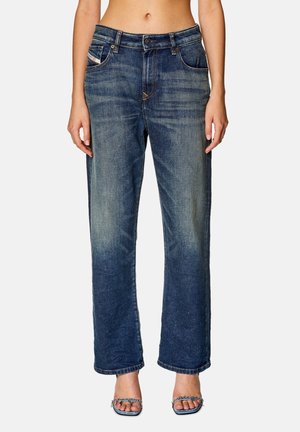 Femme portant un jean bleu taille haute, coupe droite avec un effet délavé et des sandales à talons ouvertes, debout devant un fond uni.