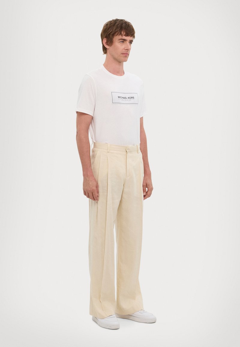 White crew-neck T-shirt with black "Michael Kors" logo, paired with beige wide-leg trousers and white sneakers. Simple design and light texture.