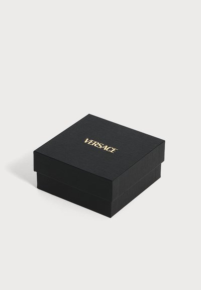 Μαύρο υφασμένο τετράγωνο κουτί με χρυσλό λογότυπο Versace στο κέντρο του καπακιού, τοποθετημένο σε λευκή επιφάνεια.