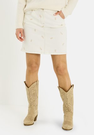 Femme portant une mini-jupe blanche avec des détails brodés subtils, des bottes de cowboy en daim beige et un pull crème avec la main dans la poche.